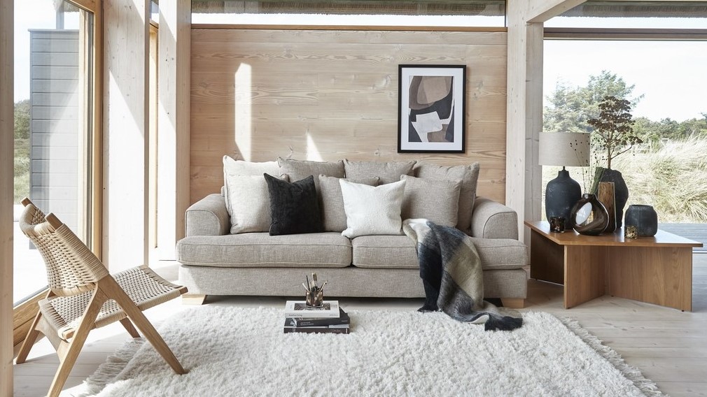 Ljus soffa med kuddar framför trävägg och fönster