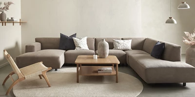 U-soffa i gråaktig färg. Framför står en fåtölj i rotting och ett soffbord i trä