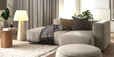 Ljus soffa med open end stående i ett ljust vardagsrum med golvlampa, puff och vit matta.