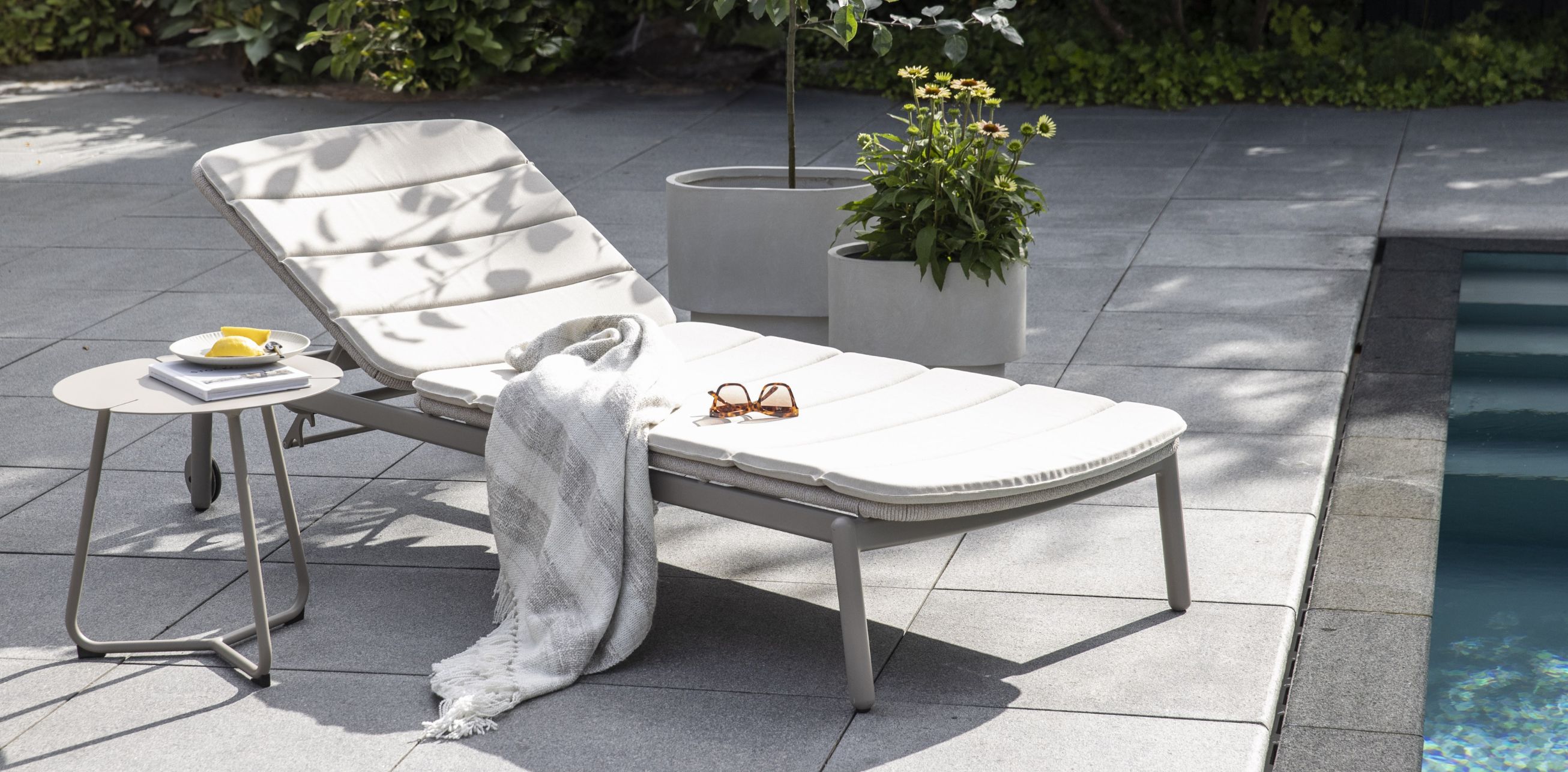 En bild av en vit solstol vid en pool med en filt, en bok och solglasögon på stolen. Bredvid stolen står ett bord med snacks och en kopp.