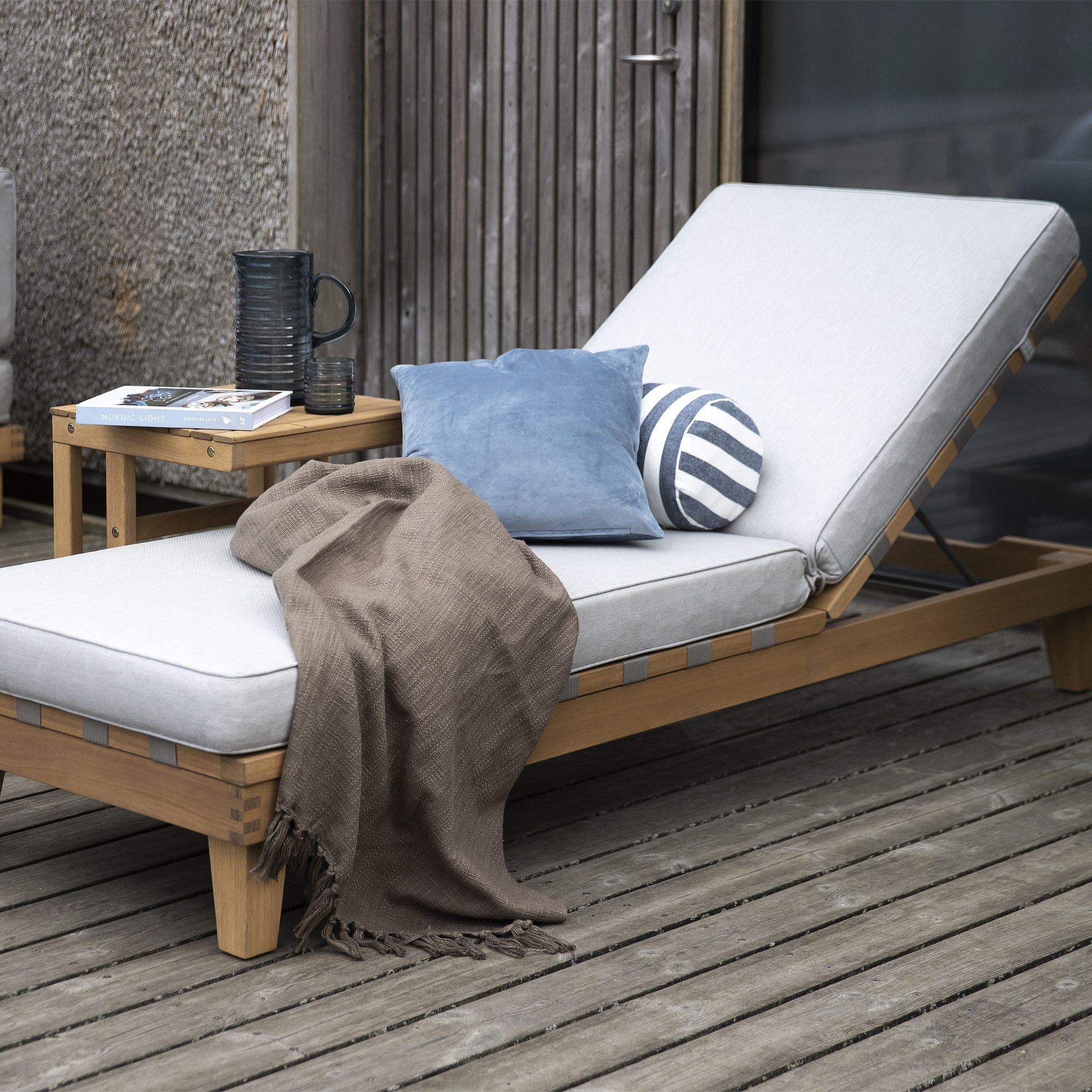 Träsolstol på en terrass med blå kudde och brunt täcke, bredvid kaffe och böcker.