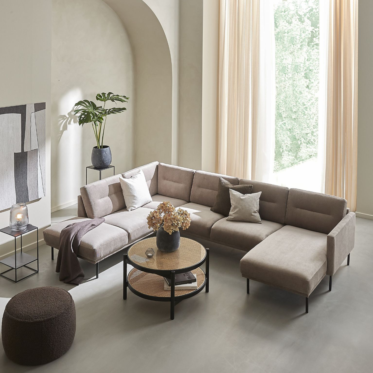 Helt vardagsrum sett ovanifrån. I rummet finns en u-soffa, ett runt soffbord med fransk rotting och svart ram, en brun puff samt några vaser och en stor tavla.