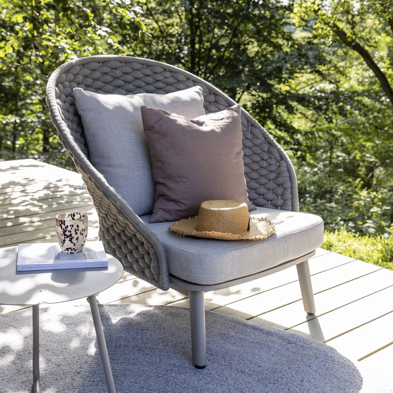 Modern böjd stol med tjocka grå kuddar och ett grovt vävt ryggstöd på en terrass med bord och kopp.