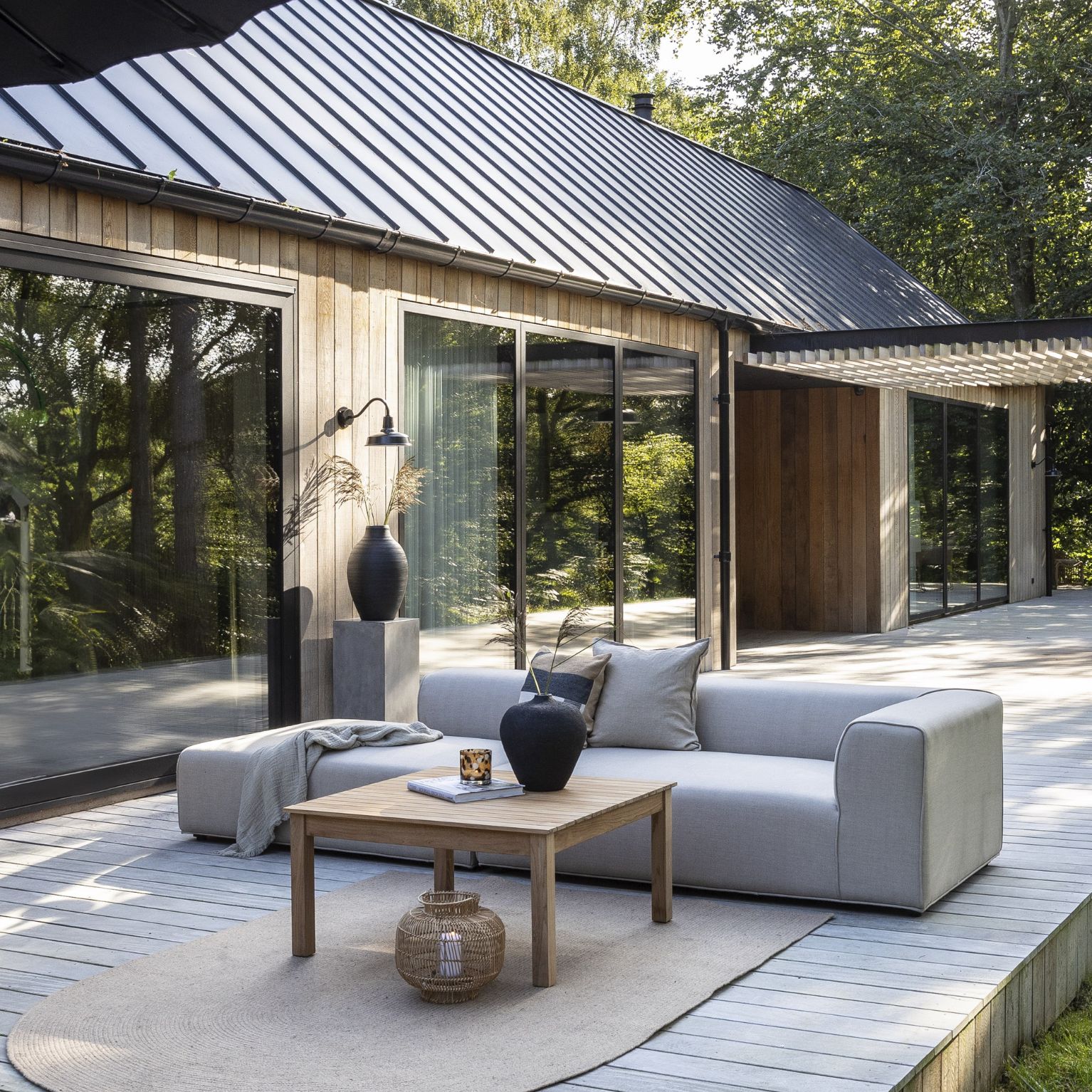 Utomhus vardagsrum med modern soffa och träbord framför ett hus med stora fönster och träklädsel.