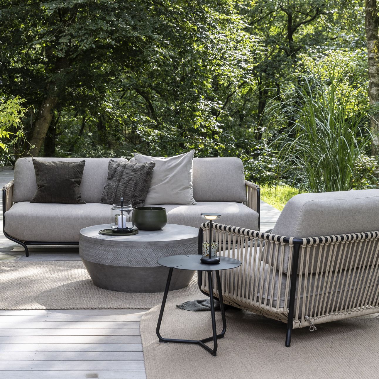 Modernt utomhus sittområde med grå soffor, runt bord och vävt ryggstöd på sofforna, omgivet av gröna växter.