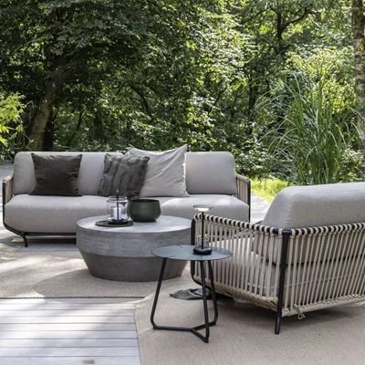 Modernt utomhus sittområde med grå soffor, runt bord och vävt ryggstöd på sofforna, omgivet av gröna växter.