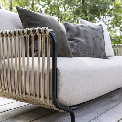 Detalj av en utomhussoffa med vävt ryggstöd och grå kuddar på en träterrass omgiven av träd.