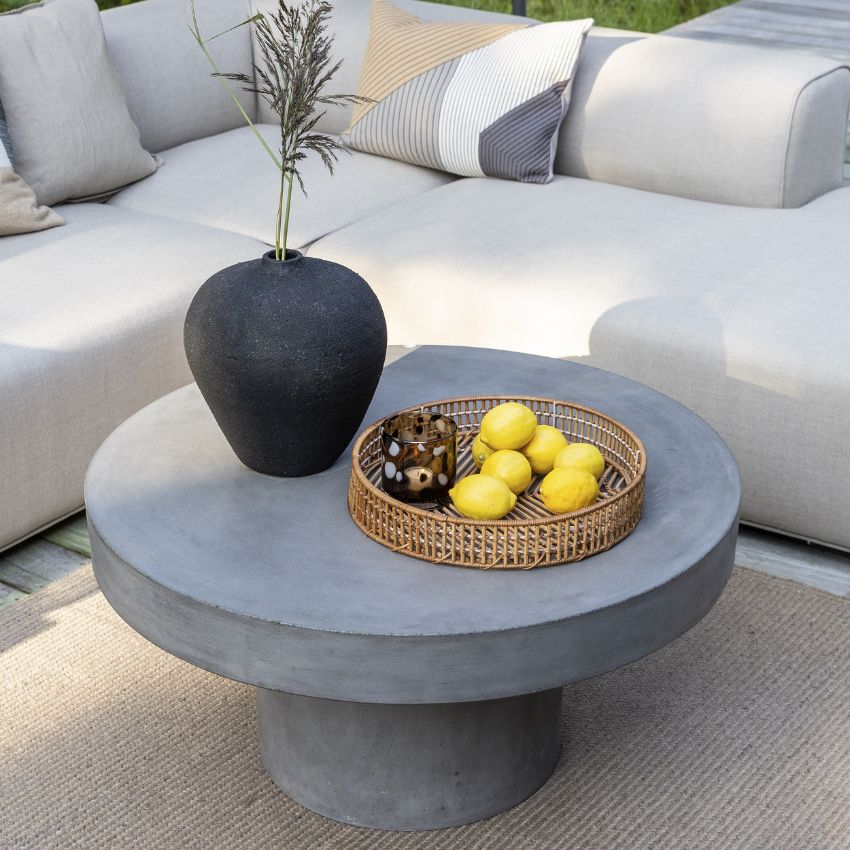 Modernt grått betongbord med en svart vas, korgfat med frukt, omgivet av mjuka vita soffor.