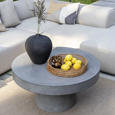 Modernt grått betongbord med en svart vas, korgfat med frukt, omgivet av mjuka vita soffor.