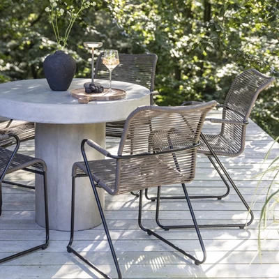 Betongbord med svart vas och glas på terrassen omgiven av vävda stolar och grön vegetation.