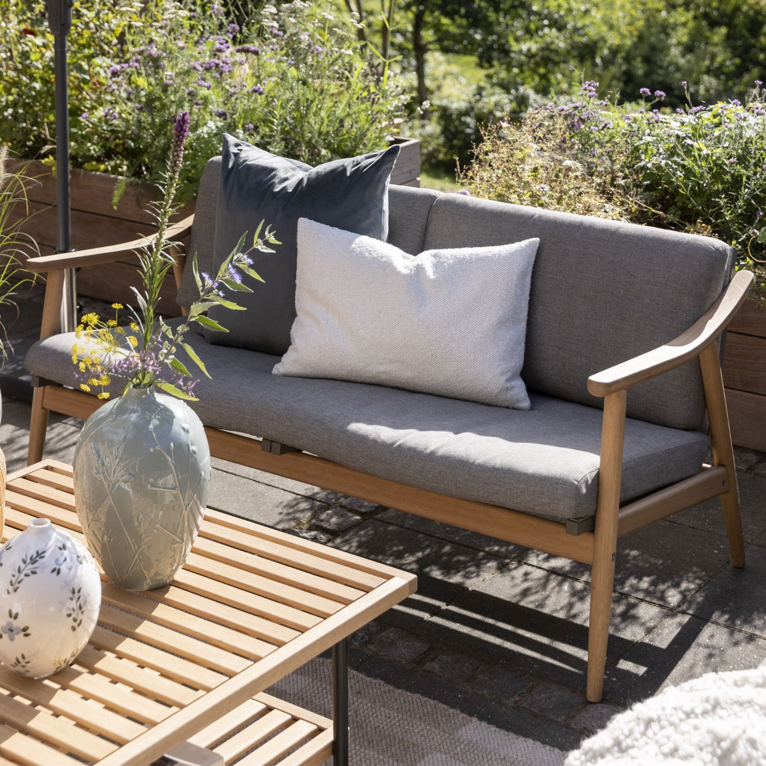 Utomhussoffa med grå kuddar och träram, dekorerad med en vas på ett träbord.