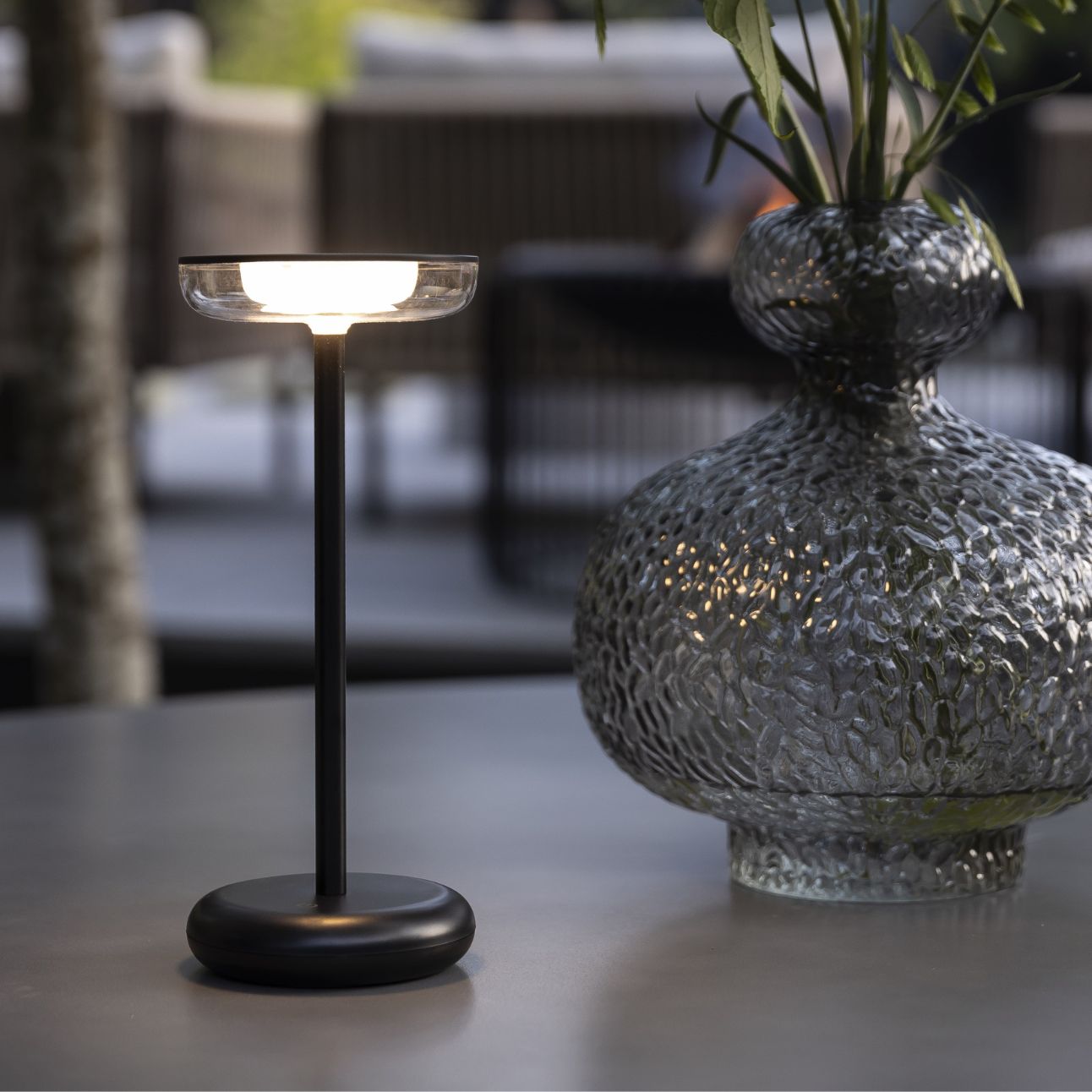 Modern bordslampa på ett terrassbord bredvid en dekorativ glasvas med växter.