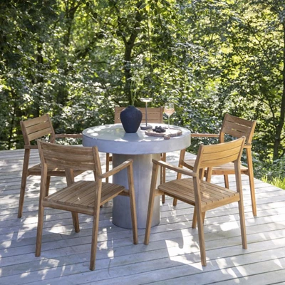 Runt utomhus matbord med trästolar, placerat på en trädterrass omgiven av träd.