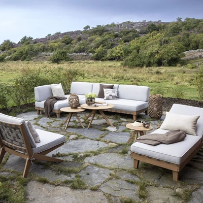 Utomhus vardagsrum med grå soffor och träbord på en stenlagd terrass med utsikt över ett öppet landskap.