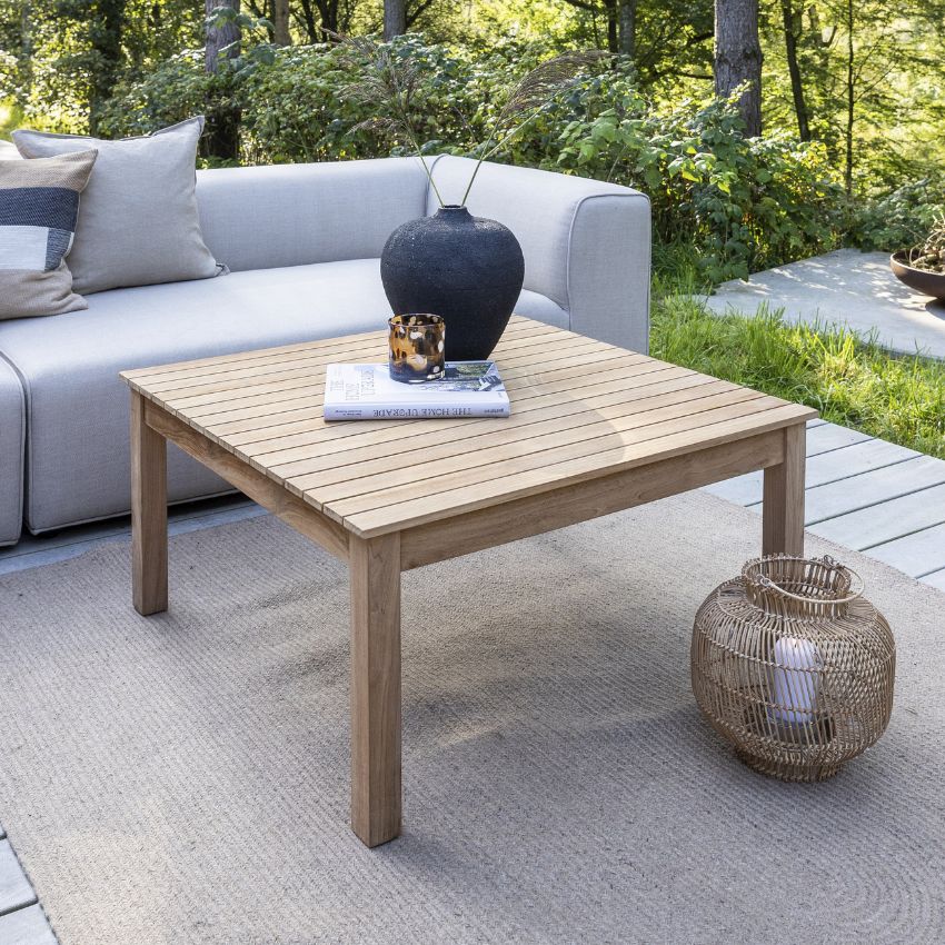 Modernt utomhus sittområde med ett träbord, en svart vas, en bok, och en korg, omgiven av en soffa och frodig växtlighet.