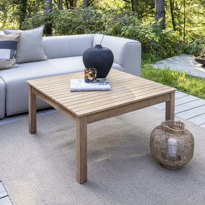 Modernt utomhus sittområde med ett träbord, en svart vas, en bok, och en korg, omgiven av en soffa och frodig växtlighet.