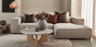 Elegant beige hörnsoffa med prydnadskuddar i varma nyanser. Ett runt marmorbord med träben och en vas med torkade blommor kompletterar inredningen