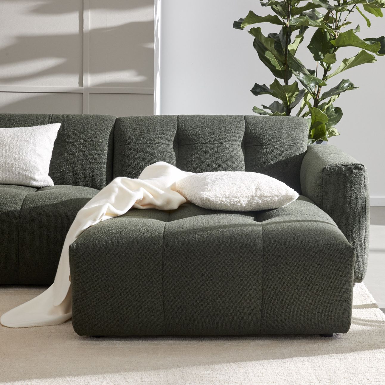 Stilren mörkgrön soffa med quiltad stoppning, en vit kudde och en filt, omgiven av ett ljust rum med en stor växt