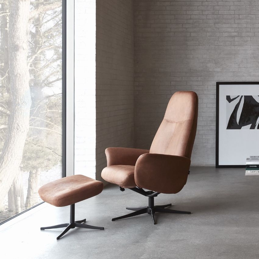 Bekväm brun fåtölj med matchande fotpall placerad i ett minimalistiskt rum med stora fönster och betongväggar
