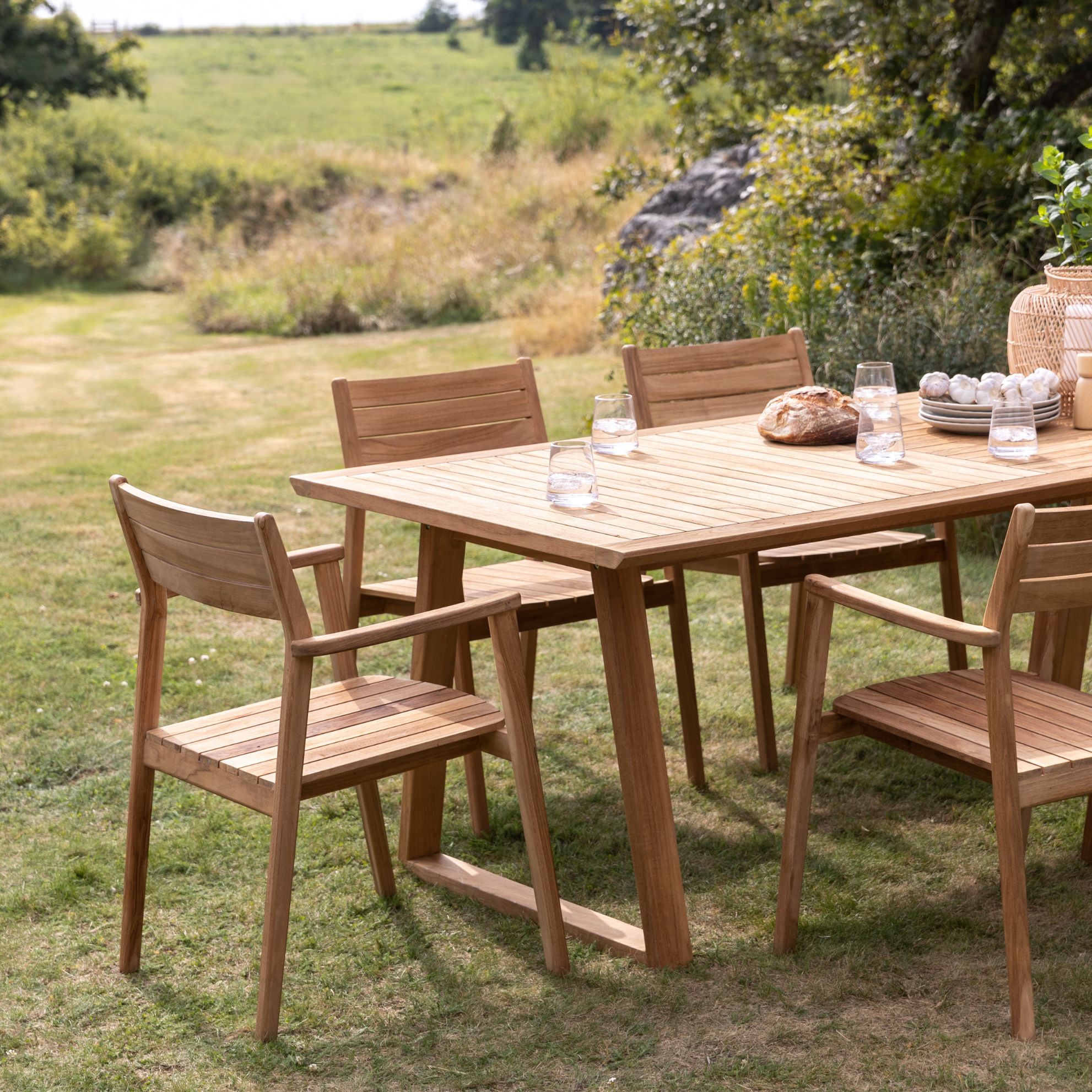 Utomhus trämatbord med stolar på en gräsmatta med kuperat landskap i bakgrunden.