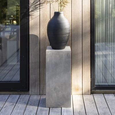 Piedestal i fibercement med en svart vas med blommor i.