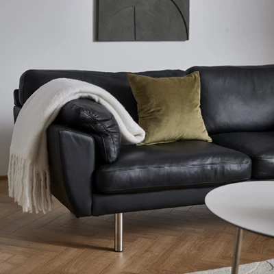 Sort lædersofa med gullig pyntepude og hvidt tæppe hængende på armlæn