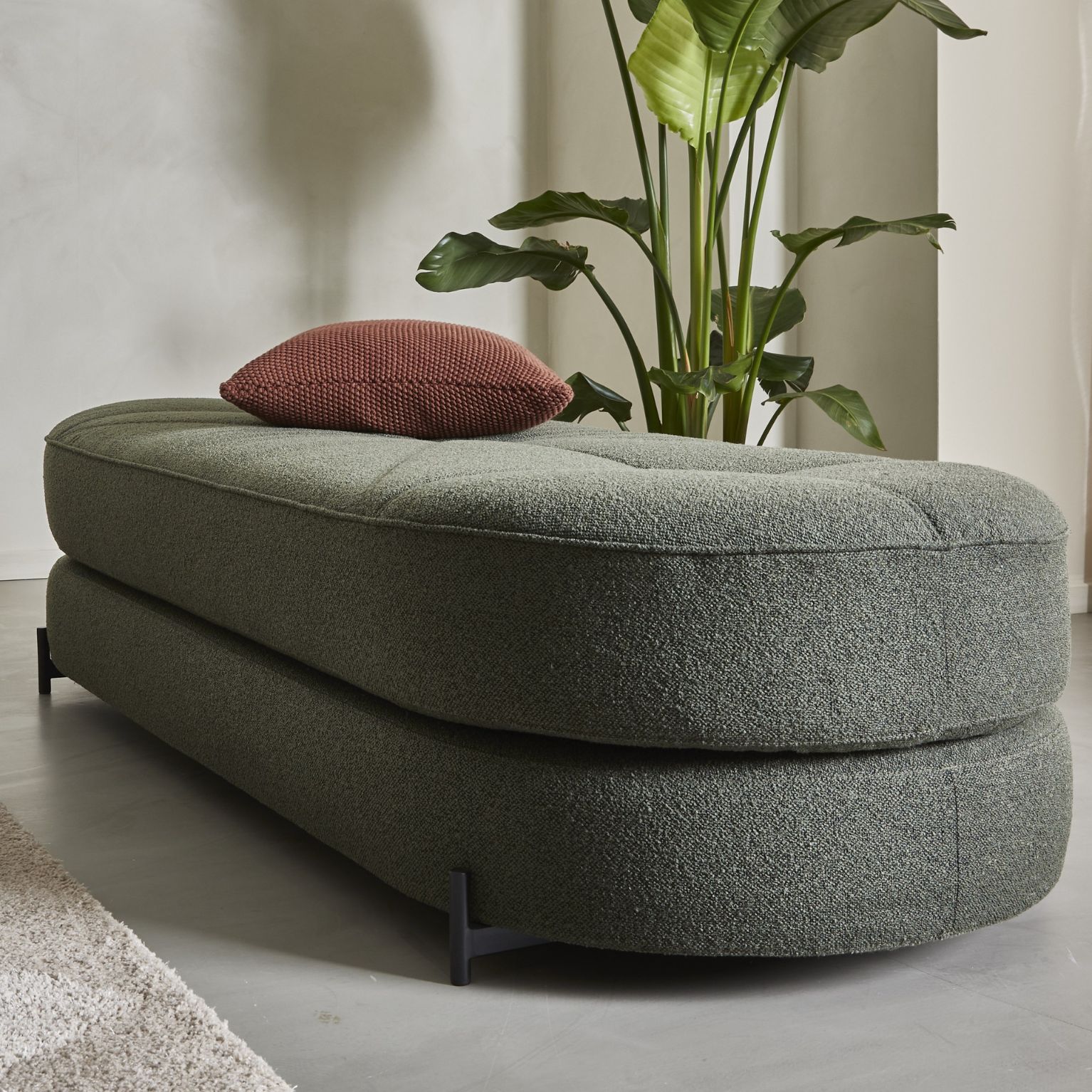 Modern och stilfull dagbädd i mörkgrönt tyg med flera lager och en mörkröd kudde ovanpå. Bakom dagbädden står en stor grön växt som tillför en frisk atmosfär till rummet.