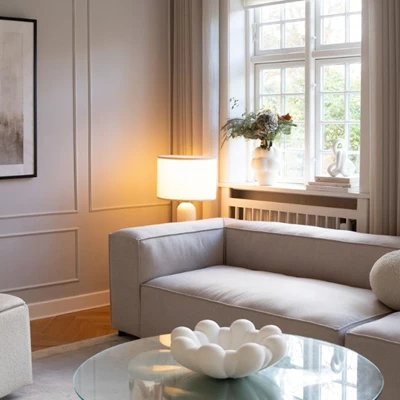 Inblick i ett mysigt vardagsrum i vita och beige nyanser, där det står en modulsoffa, soffbord med en vas ovanpå, en bordslampa i hörnet, samt stilfulla gardiner i sand