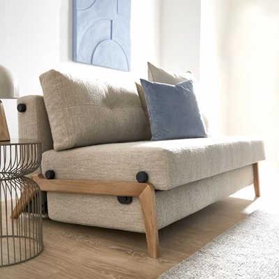 Bäddsoffa med träram i grå textil som står på ett trägolv med kuddar på sitsarna