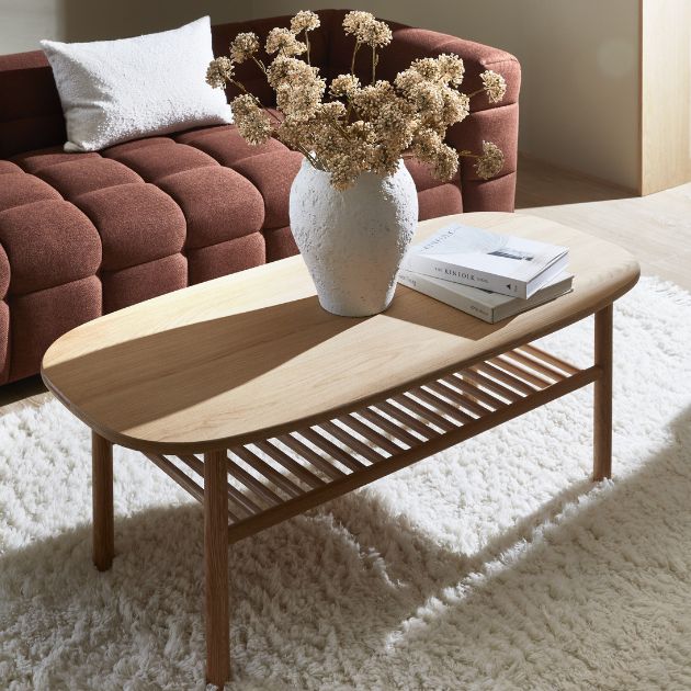 Ljust soffbord i trä med en vit keramisk vas, torkade blommor och en bok, placerat framför en brun quiltad soffa på en mjuk vit matta