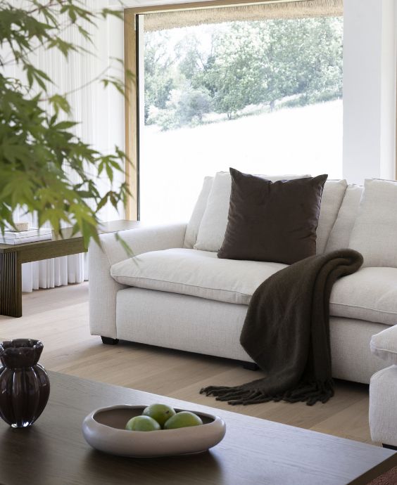 Vit soffa med en brun kudde och filt placerad framför ett stort fönster, med en skål med gröna äpplen på soffborde
