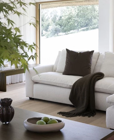 Vit soffa med en brun kudde och filt placerad framför ett stort fönster, med en skål med gröna äpplen på soffborde