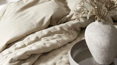 Säng med beige överkast och vas med torkade blommor på nattduksbordet