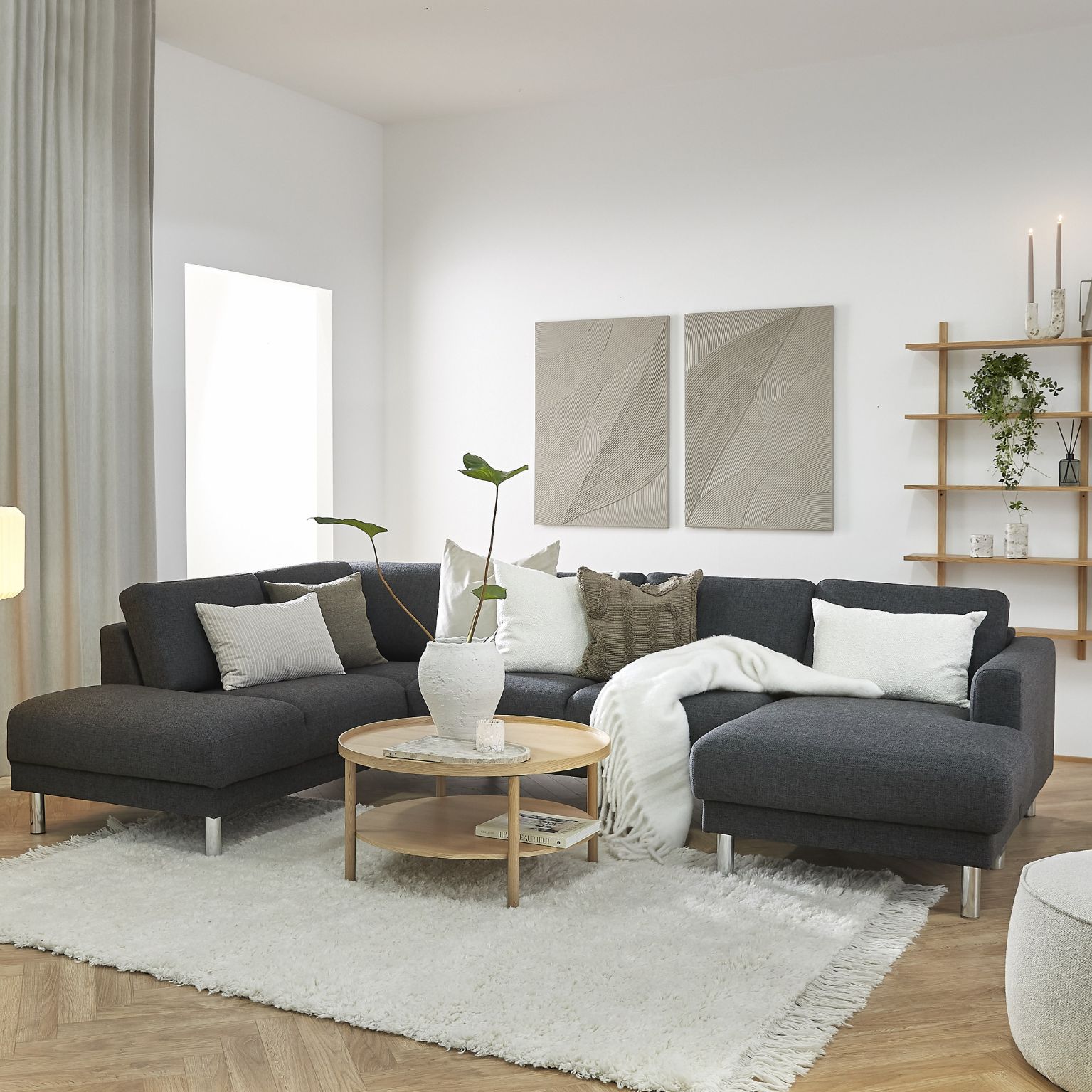 Hörnsoffa med chaise longue i mörkt tyg stående i vardagsrumsinredning. Den står ovanpå en vit matta tillsammans med ett runt soffbord i trä