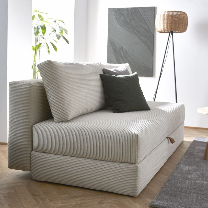 Bäddsoffa i sammetstyg, dekorerad med mörkgrå kuddar. Bakom soffan hänger en grå abstrakt konstplansch, och bredvid står en flätad golvlampa med tre ben