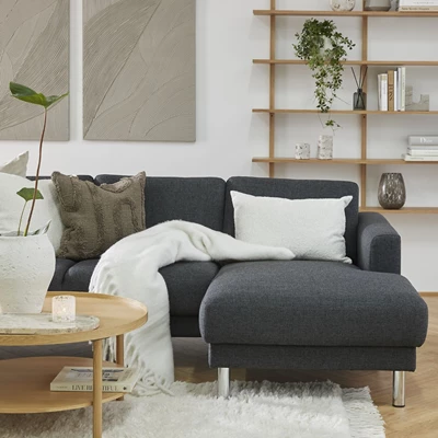 Schäslongsoffa i mörkgrå klädsel på vit matta tillsammans med ett soffbord i trä. Bakom hänger två canvasmålningar och en vägghylla i trä.