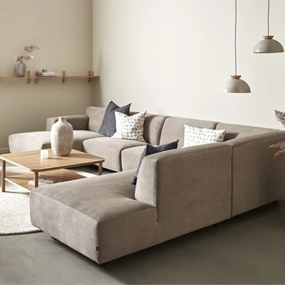 U-soffa i gråbrun klädsel på betonggolv. På väggen hänger en trähylla och på golvet ligger en vit matta med ett soffbord ovanpå.