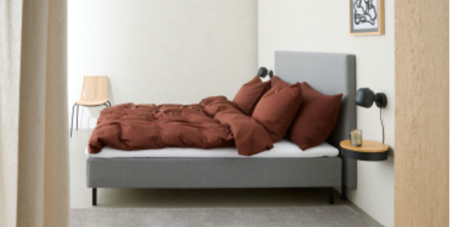 En boxmadrass i grått sedd från sidan. Sängen är bäddad med rött sängkläder.