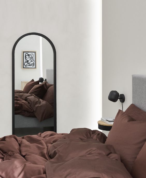 Säng med rött sänglinne stående bredvid en avlång spegel med svart kant hängande på väggen