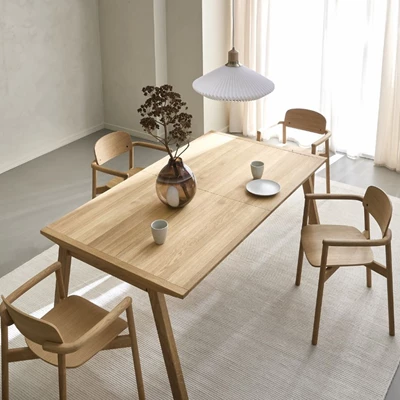 En modern matsal med ett långt träbord, fyra trästolar, en vit pendellampa och en vas med en torkad växt på bordet i ett ljust rum med beige väggar och vita gardiner.