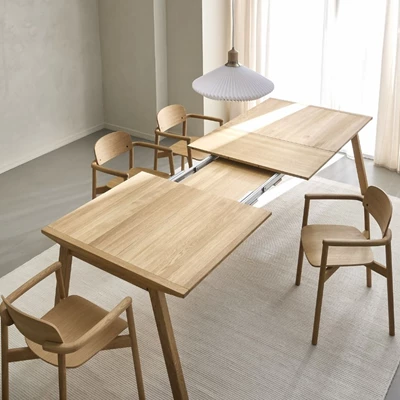 Ett matbord i trä som visas under utdragsprocessen med dolda iläggsskivor, omringat av fyra trästolar under en vit pendellampa i ett minimalistiskt designat rum.