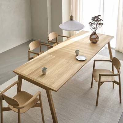 Ett fullt utdraget långt träbord i en modern matsal, flankerat av fyra trästolar och dekorerat med en vas och torkade växter under en stor vit pendellampa i ett rum med mjukt naturligt ljus.