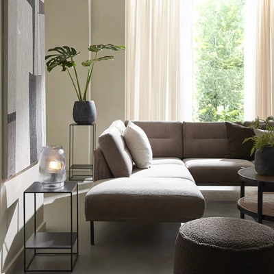 Utdrag av vardagsrum med en soffa och en puff i grått tyg i centrum. Runtomkring finns bland annat två sidobord med en lampa respektive en planta i svart kruka.