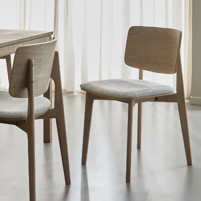 Två stolar med trästomme och stoppade säten. Stolarna har böjt ryggstöd och står vid ett träbord. Bakom finns gardiner