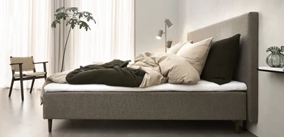 Design uden navn - 2025-01-16T135708.646.jpg Nocturne resårmadrass med rörigt sängkläder sett från sidan. Sängen är grå och med sänggavel.