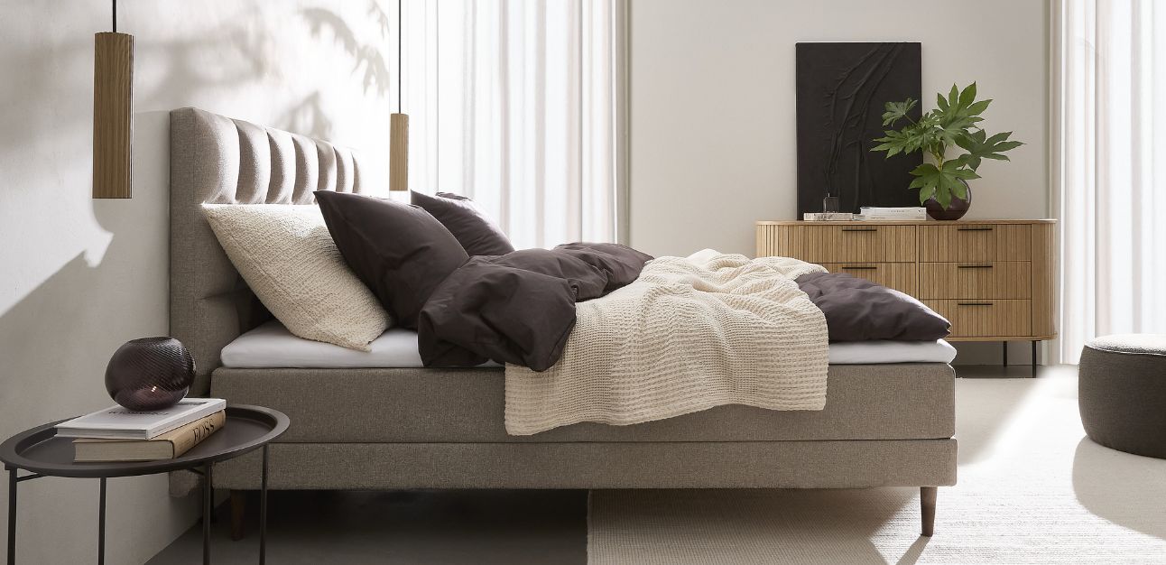 Grålig justerbar säng stående i sovrummet. Sängen har ett rutigt sänggavel, och runt om finns ett nattduksbord, en pall, en byrå samt två eleganta pendellampor på vardera sida om sängen.