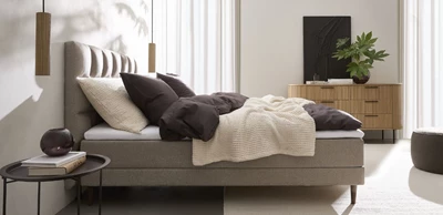 Design uden navn - 2025-01-16T141853.057.jpg Grålig justerbar säng stående i sovrummet. Sängen har ett rutigt sänggavel, och runt om finns ett nattduksbord, en pall, en byrå samt två eleganta pendellampor på vardera sida om sängen.