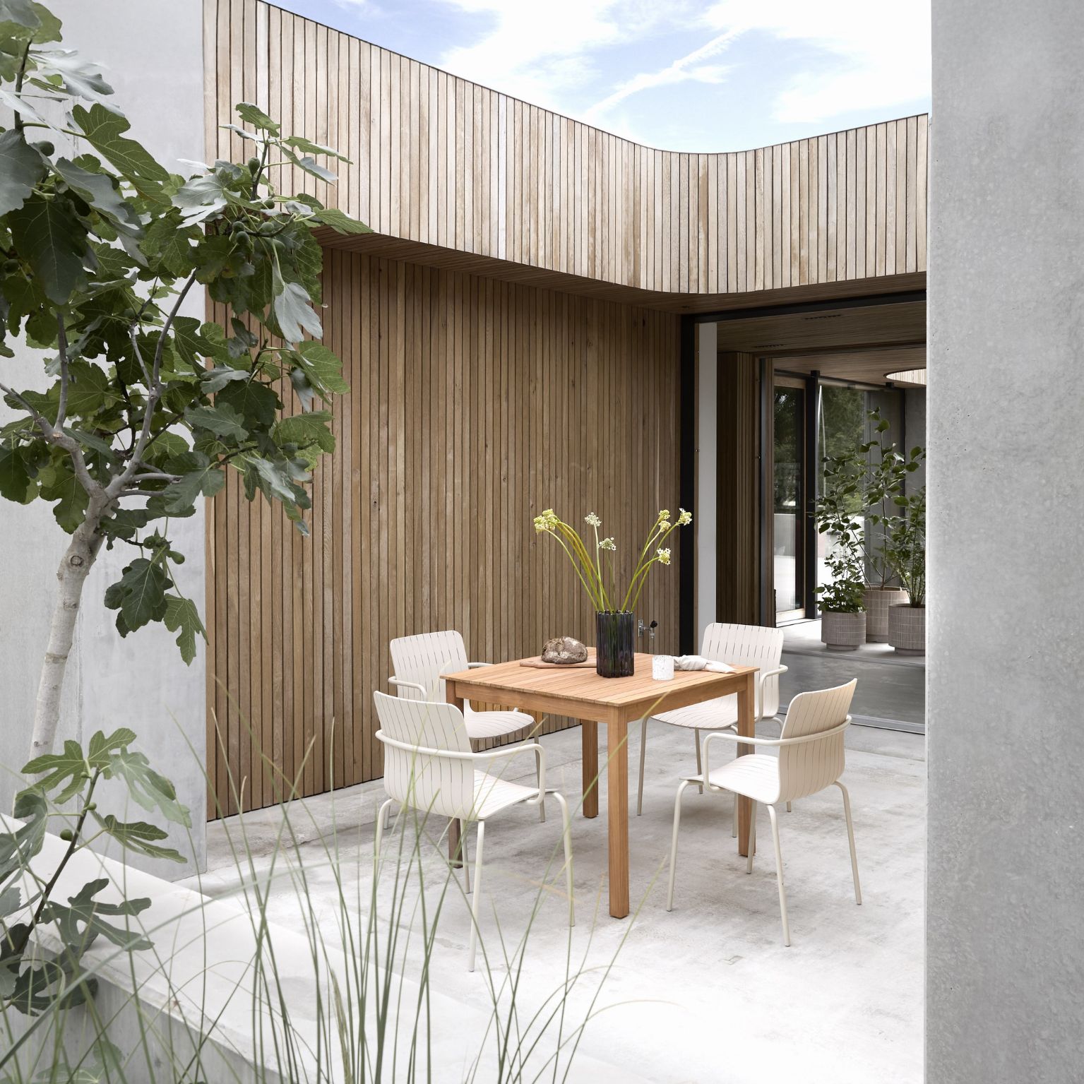 Utomhusmatplats med ett fyrkantigt träbord omgivet av fyra ljusa stolar med ribbat design. Bordet är dekorerat med en svart vas med höga, gula blommor samt små dekorationer. Omgivningarna har en modern och minimalistisk stil med träbeklädda väggar, betongytor och gröna växter som skapar en harmonisk atmosfär.