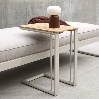 Litet sidobord med träskiva och ljust metallunderrede placerat vid en stoppad bänk. Bordet har en modern och funktionell design där underredet kan skjutas in under bänken. På bordet står en liten vit bordslampa och en brun vas.