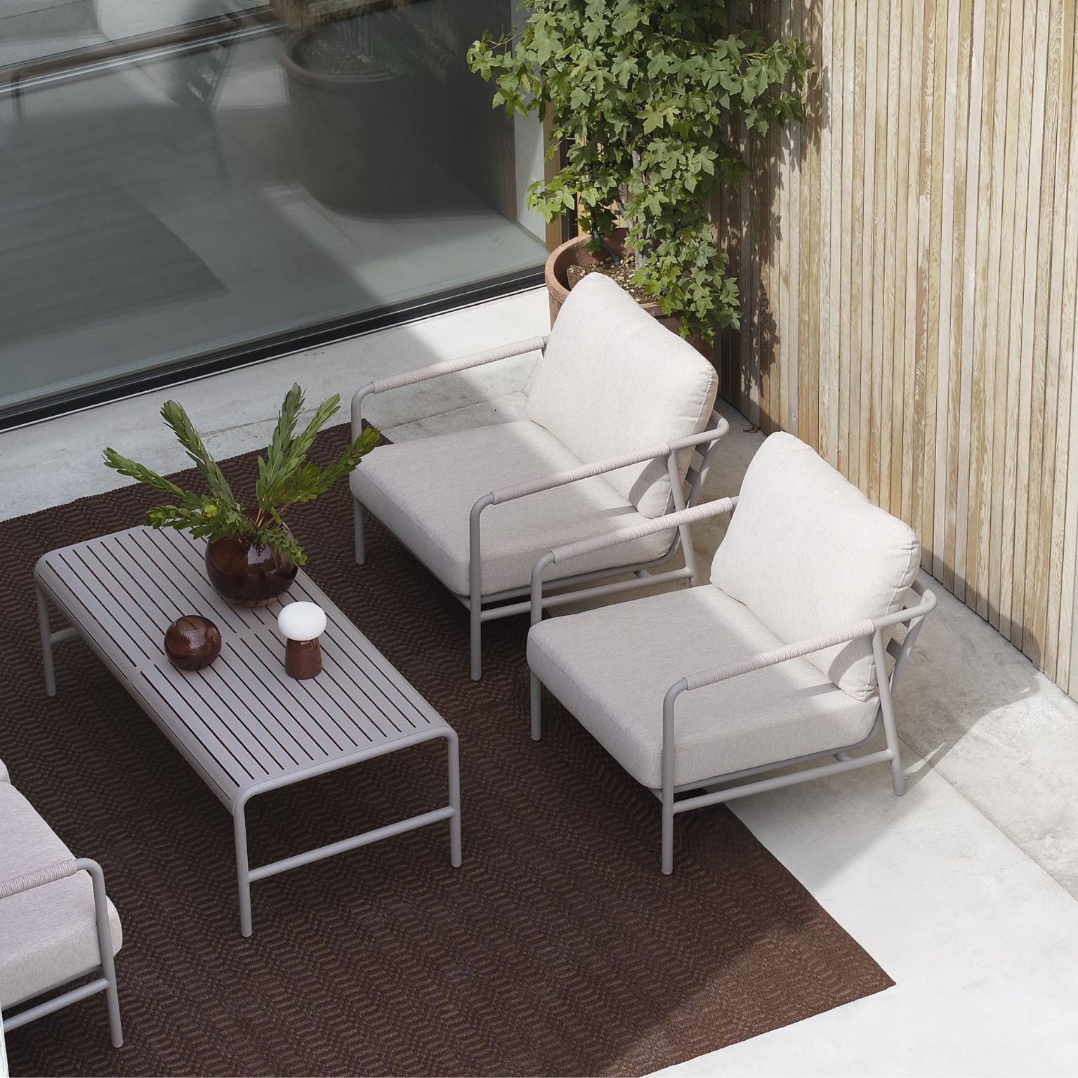 Utomhusloungeområde med två ljusa loungestolar med metallunderrede och mjuka dynor, placerade runt ett rektangulärt soffbord med lamelldesign. Området är inrett med en brun utomhusmatta och omgivet av en modern kombination av träbeklädda väggar, betong och gröna växter, vilket skapar en stilren och avslappnad atmosfär.
